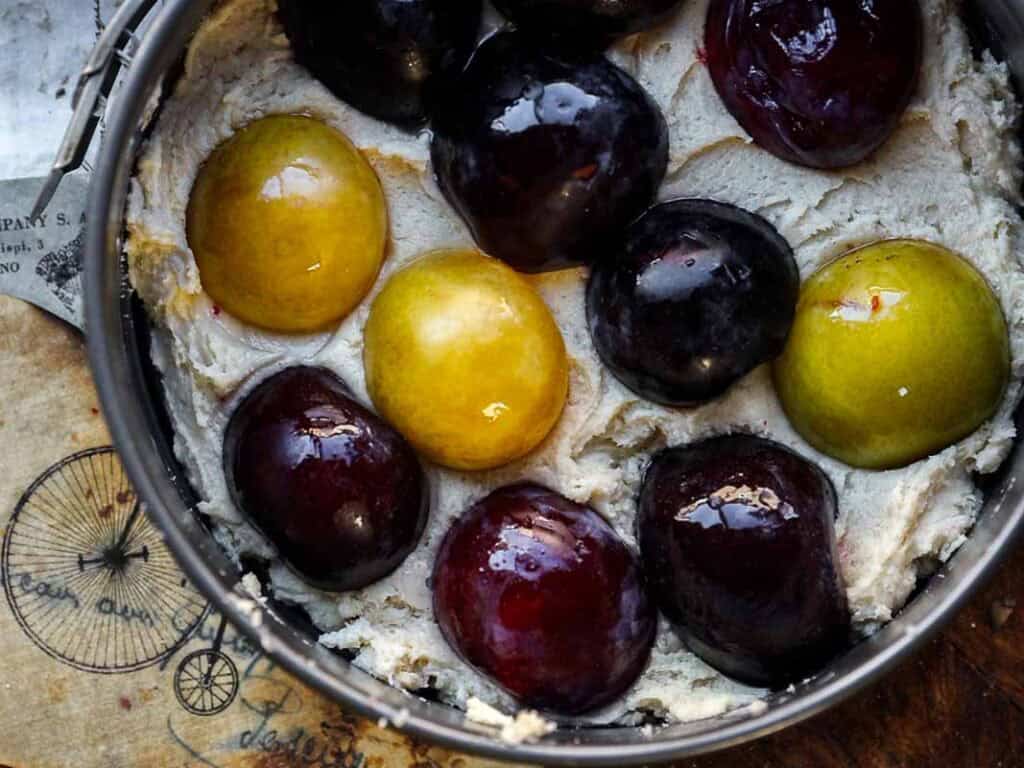 A baking pan filled with creamy cake batter, topped with whole yellow and dark purple plums arranged in a circle, ready to be baked. The pan sits on a wooden surface with printed text and a bicycle illustration.